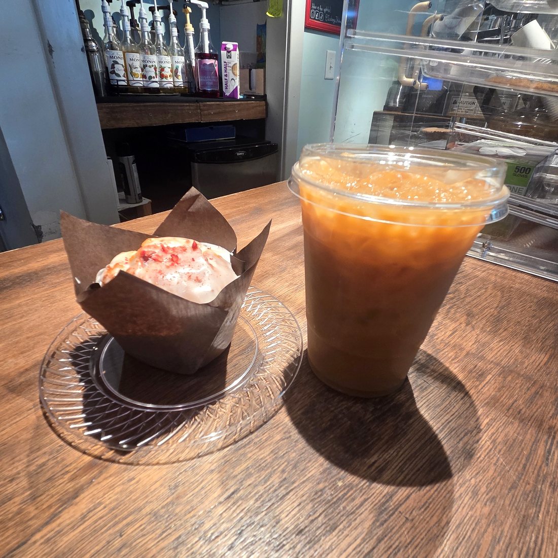 An iced coffee and a glazed muffin on the wooden counter, with the syrup rack and pastry case behind.